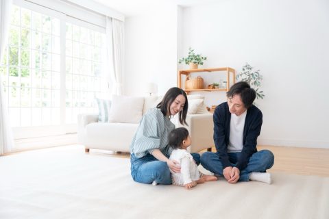 明るく開放的なリビングの施工事例。大きな窓から自然光が降り注ぐ白い空間で、夫婦と赤ちゃんが笑顔でくつろぐ。