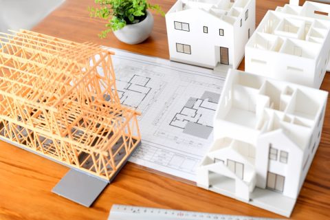 木造建築の骨組み模型、複数の白い住宅模型、設計図面が並べられている。注文住宅の建築設計や家づくりの計画段階を示す施工事例。