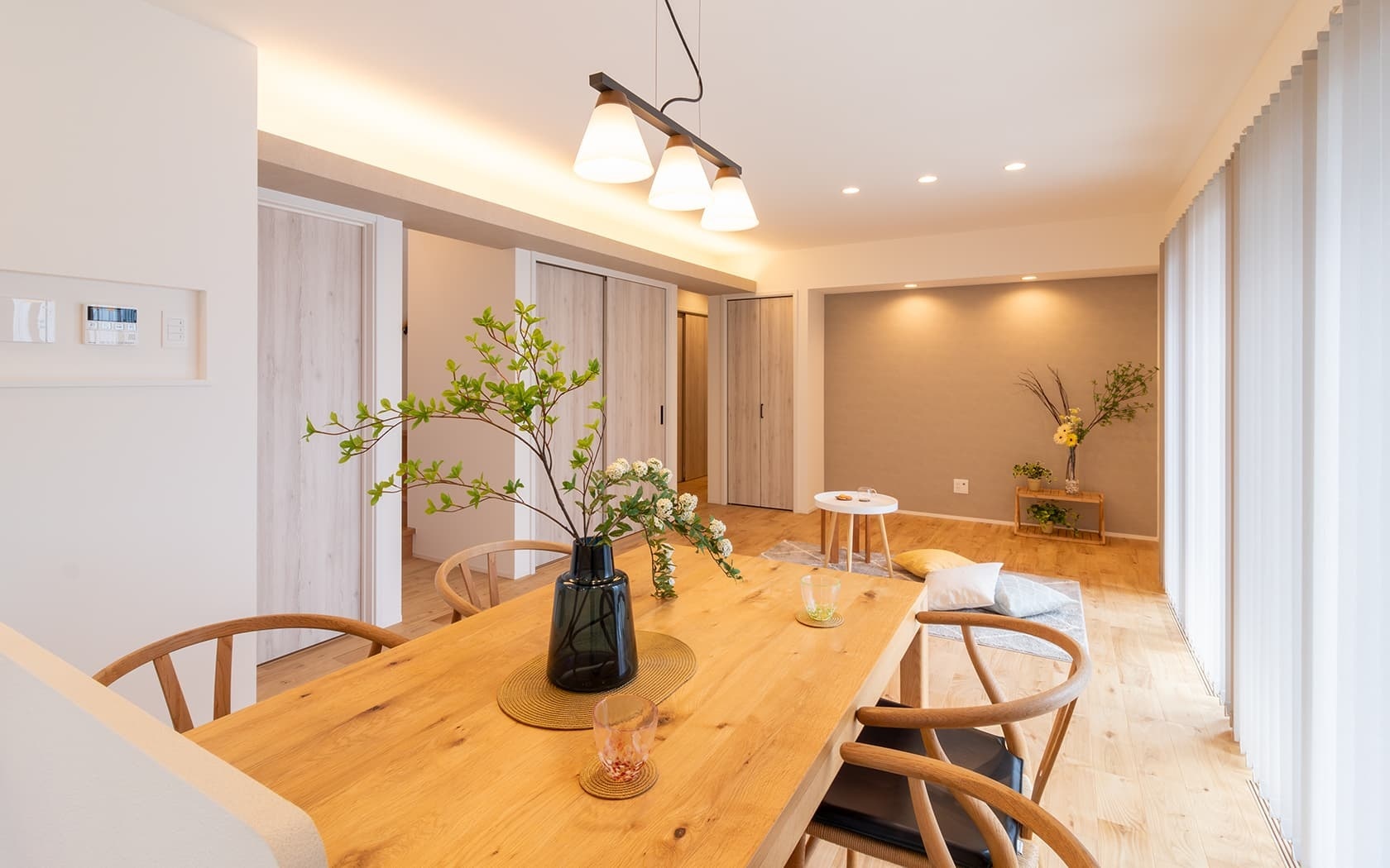 明るく開放的なダイニング・リビングルームの施工事例。無垢材の大きなダイニングテーブルに木製チェアが並び、モダンなペンダントライトが空間を照らす。奥には木目調の収納扉と、落ち着いたアクセントウォールに間接照明が配されたリビングスペースが見える。ナチュラルで温かみのあるモダンな内装デザインが特徴。