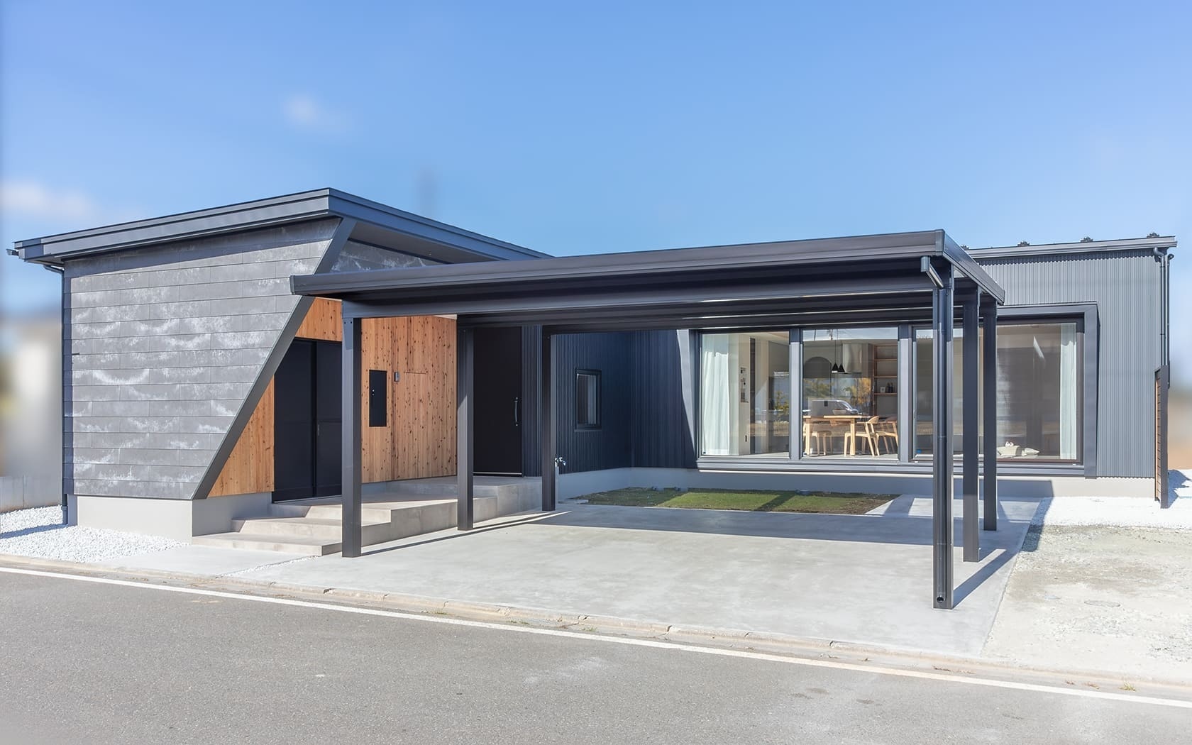 グレーのガルバリウム鋼板と木材を組み合わせたモダンな平屋住宅の全景。住宅と一体化したようなデザインの黒いカーポートが特徴で、大きな窓から明るい室内が見えるスタイリッシュな外観デザインの施工事例。