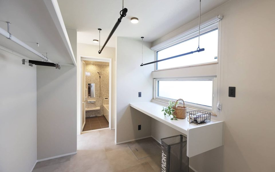 明るく広々としたランドリールームの施工事例。大きな窓から自然光が差し込み、天井には昇降式の物干し竿が複数設置されている。窓下には洗濯物を畳んだりアイロンがけに便利な白いカウンターがあり、奥には脱衣所と浴室が続く。家事動線を考慮した機能的な空間で、雨の日でも快適に室内干しができる。