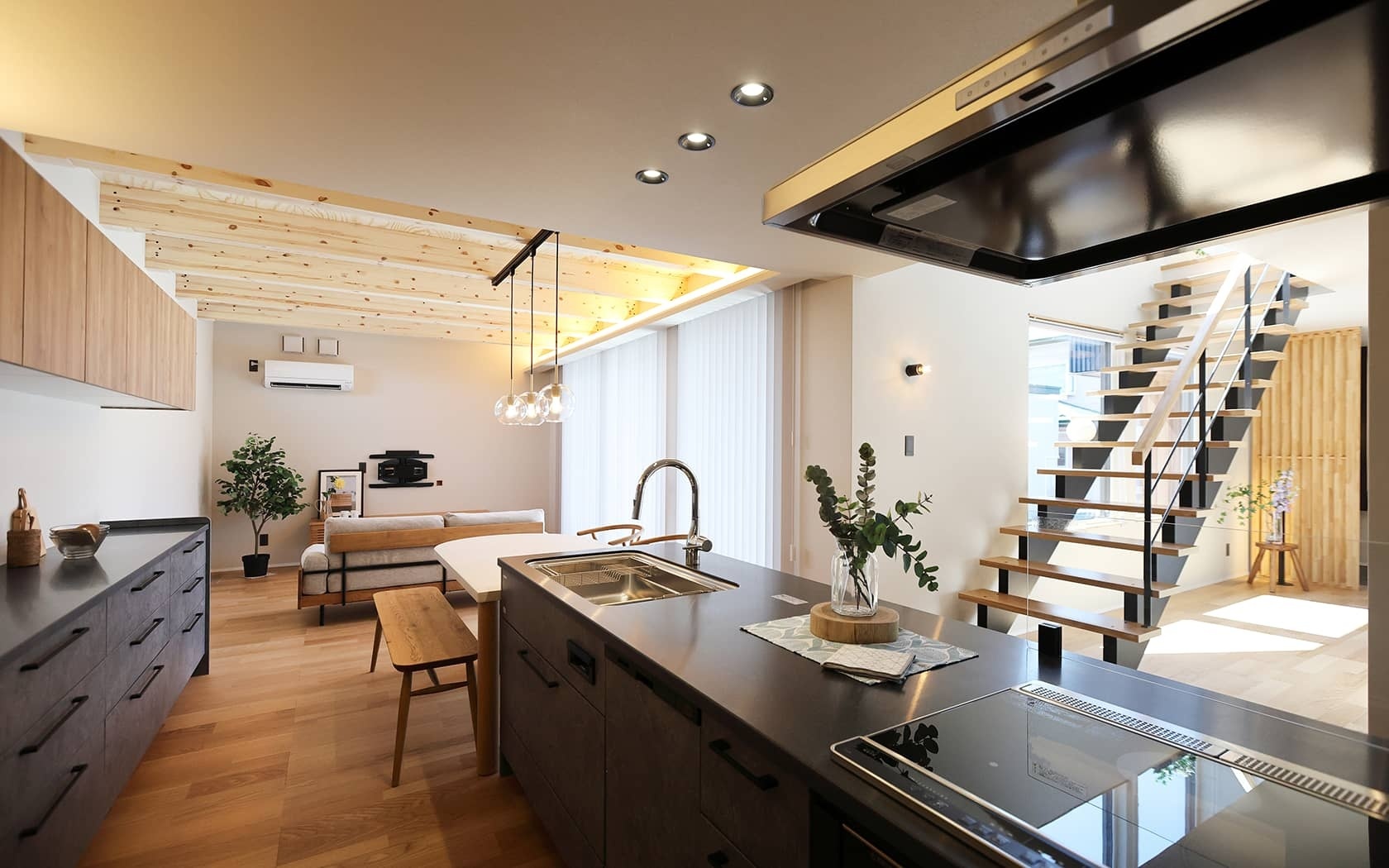 木目の天井と明るいフローリングが特徴の、モダンで開放的なリビングダイニングキッチン。ダークグレーのアイランドキッチンにはシンクとIHが備わり、隣には木製ベンチ付きのダイニングテーブル。奥にはソファのあるリビングスペースと、デザイン性の高いオープン階段が見える。