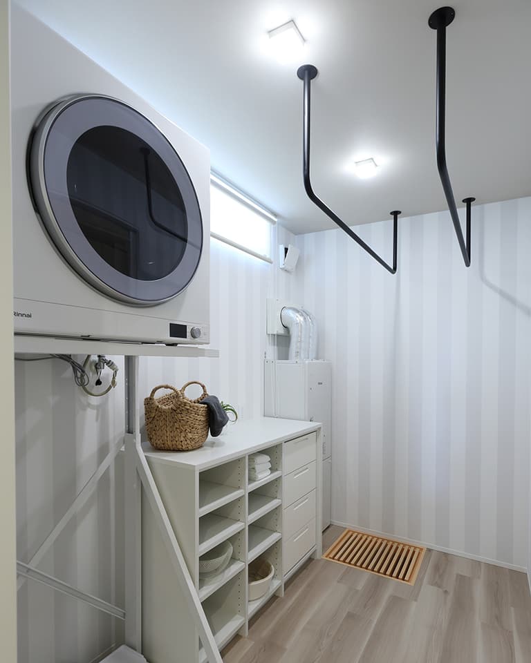 機能的でモダンなランドリールームの施工事例。ドラム式洗濯乾燥機の下に、タオルや洗剤を収納できる棚と引き出し付きキャビネットを設置。天井には黒いL字型とV字型の室内物干し竿があり、洗濯から乾燥、収納、室内干しまで完結できる効率的な家事動線を実現。白とグレーのストライプ壁紙と木目調の床が清潔感を演出。