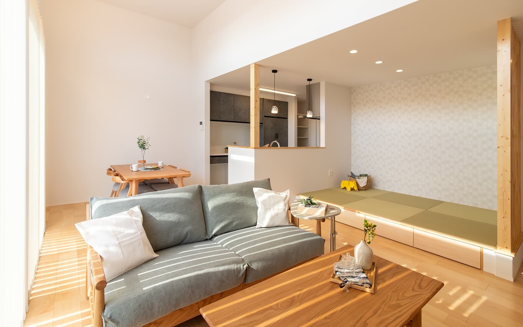 自然光が差し込む勾配天井の明るいリビングダイニングキッチンと小上がりの和室が一体となった住宅の施工事例。木製家具で統一されたナチュラルモダンな空間には、グレーのソファ、ダイニングセット、奥にモダンなキッチンが見える。収納引き出しと間接照明付きの小上がり和室が、くつろぎの場を提供する。