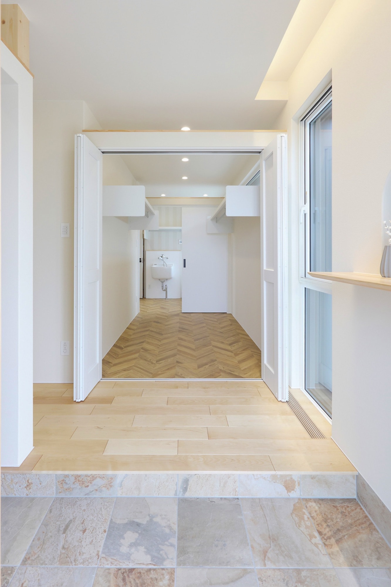 白を基調とした明るいウォークスルークローゼットの施工事例。内部には洗面台が設置され、床はヘリンボーン柄のフローリング。手前の廊下は木目調フローリング、さらに手前はベージュ系のタイル床で、異なる素材が空間を区切る。収納力と機能性を両立したモダンな内装デザイン。