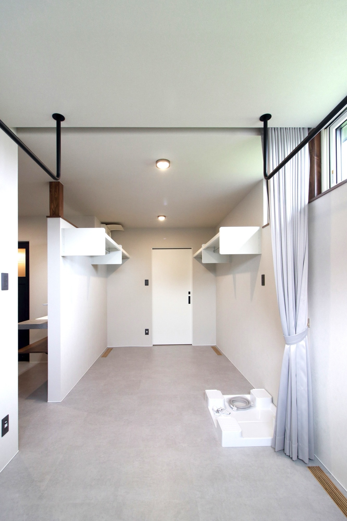 モダンなウォークスルークローゼットの施工写真。明るいグレーの床と白い壁の空間に、天井から吊り下げられた黒い室内物干しバーと白い壁付け収納棚が設置されている。右奥には洗濯パンがあり、窓には薄いグレーのカーテンがかかる。ダウンライトが明るく照らす、機能的でシンプルなデザインの空間。