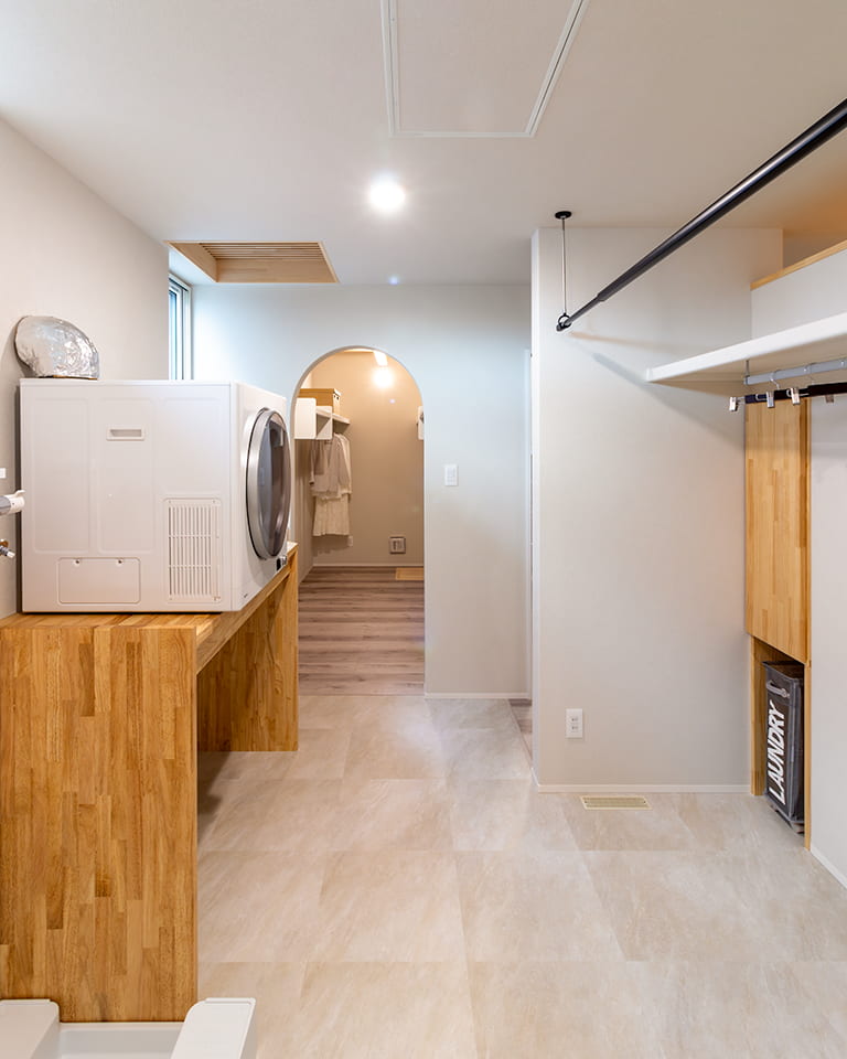 白と木目を基調とした、機能的なランドリールーム兼ウォークインクローゼット。木製カウンターにドラム式洗濯乾燥機が設置され、天井には室内干し用の物干し竿、右側には収納棚とランドリーボックスが備え付けられている。奥にはアーチ状の開口部から衣類が並ぶクローゼットが続く、家事動線を考慮した明るい空間。