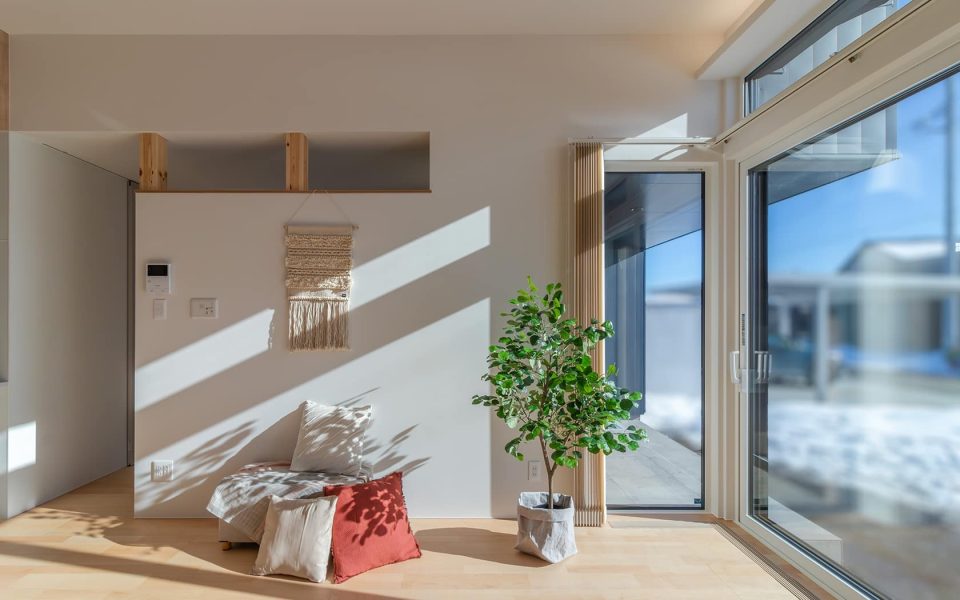 明るい日差しが差し込むモダンなリビングルーム。大きな窓からは雪景色が見え、白い壁と木製の床に光と影のコントラストが美しい。壁のタペストリーや観葉植物、クッションが置かれたベンチが、開放的で心地よい空間を演出する。