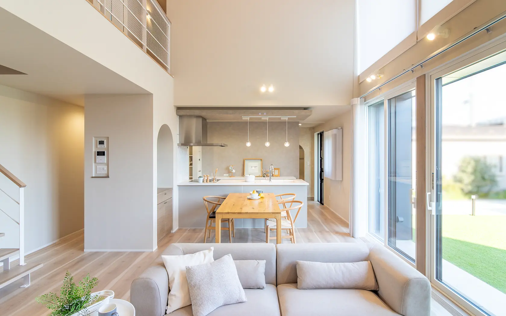 高天井と吹き抜けが開放的な広々としたリビングダイニングキッチン。明るい木目調フローリングにベージュのL字ソファ、奥にはアイランドキッチンと木製ダイニングテーブルが配置され、大開口窓から自然光が差し込むモダンでナチュラルな住宅の施工事例。