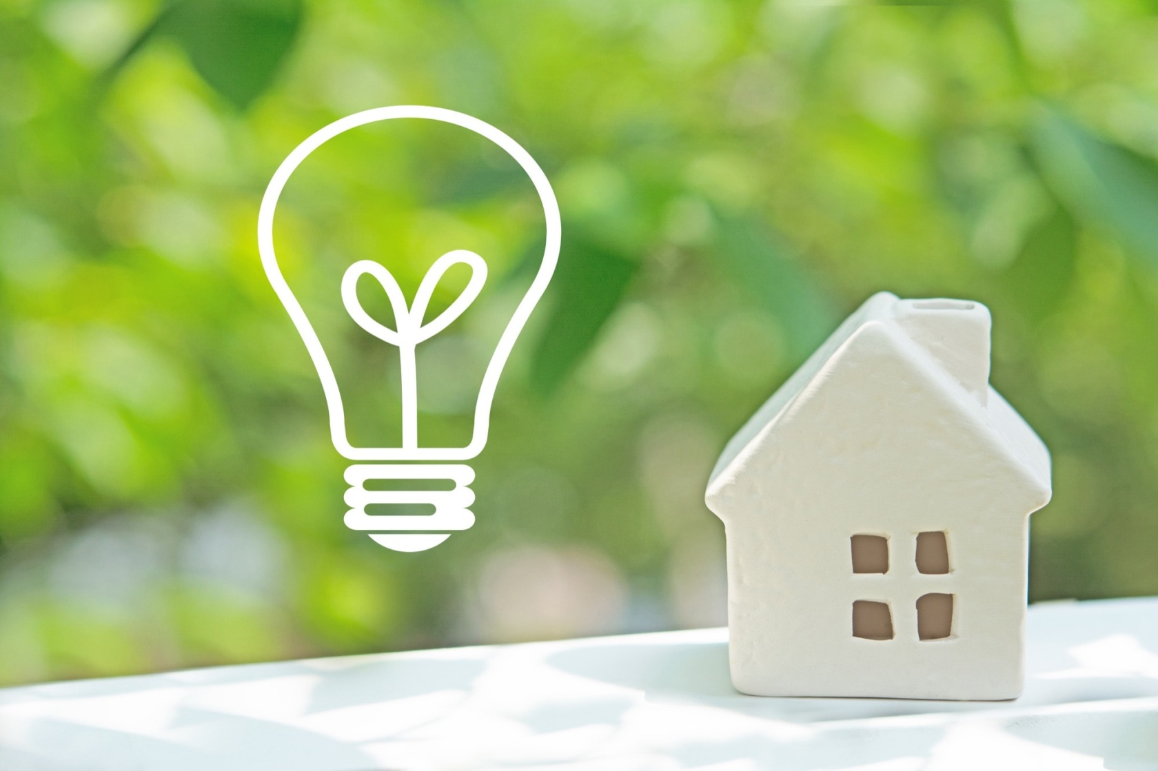 緑のぼかしを背景に、白い電球のアイコンとミニチュアの家が並ぶ。省エネ住宅、エコな暮らし、スマートハウスのアイデアを象徴。