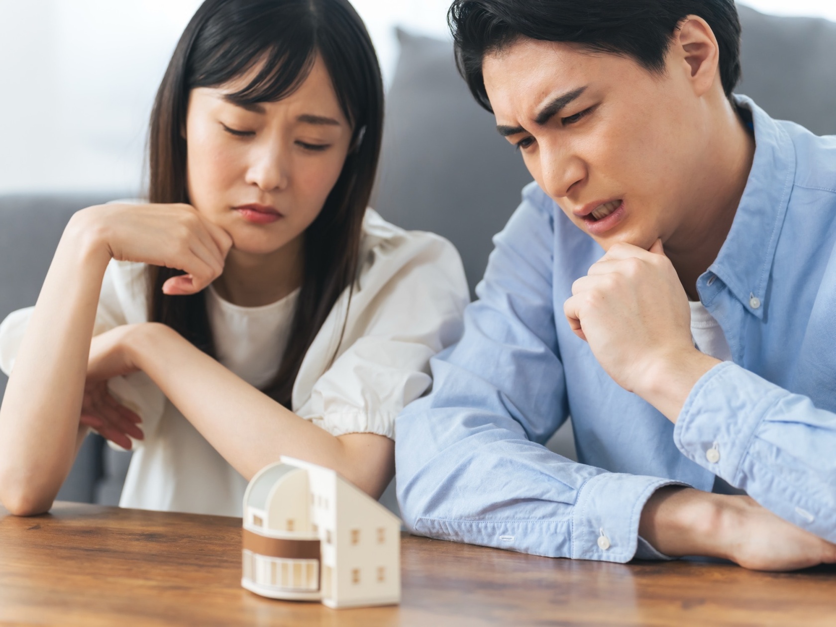 家の模型を前に深刻な表情で考え込む若い夫婦。住宅ローンや不動産購入、将来設計に関する夫婦の悩みや相談を象徴。