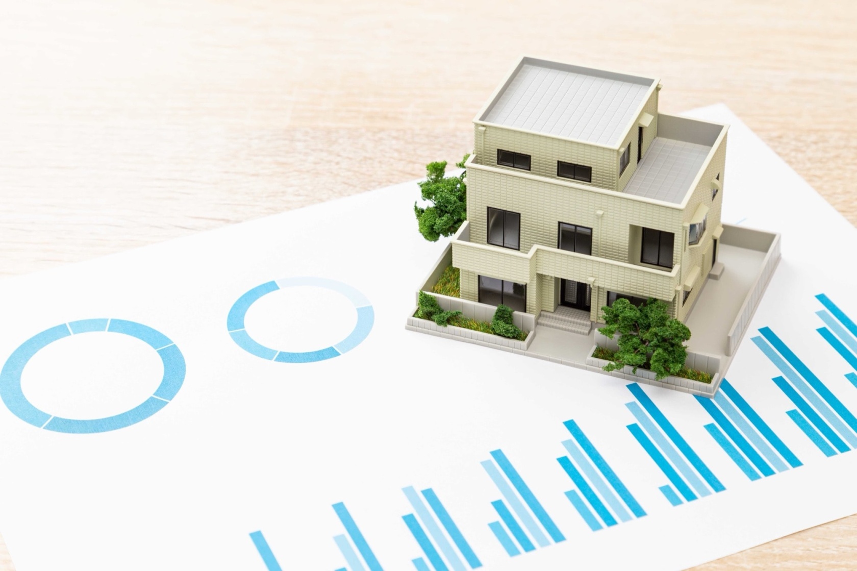 青いグラフが印刷された書類の上に置かれた一軒家のミニチュア模型。不動産投資、住宅ローン、資産運用、住宅購入のデータ分析や計画。