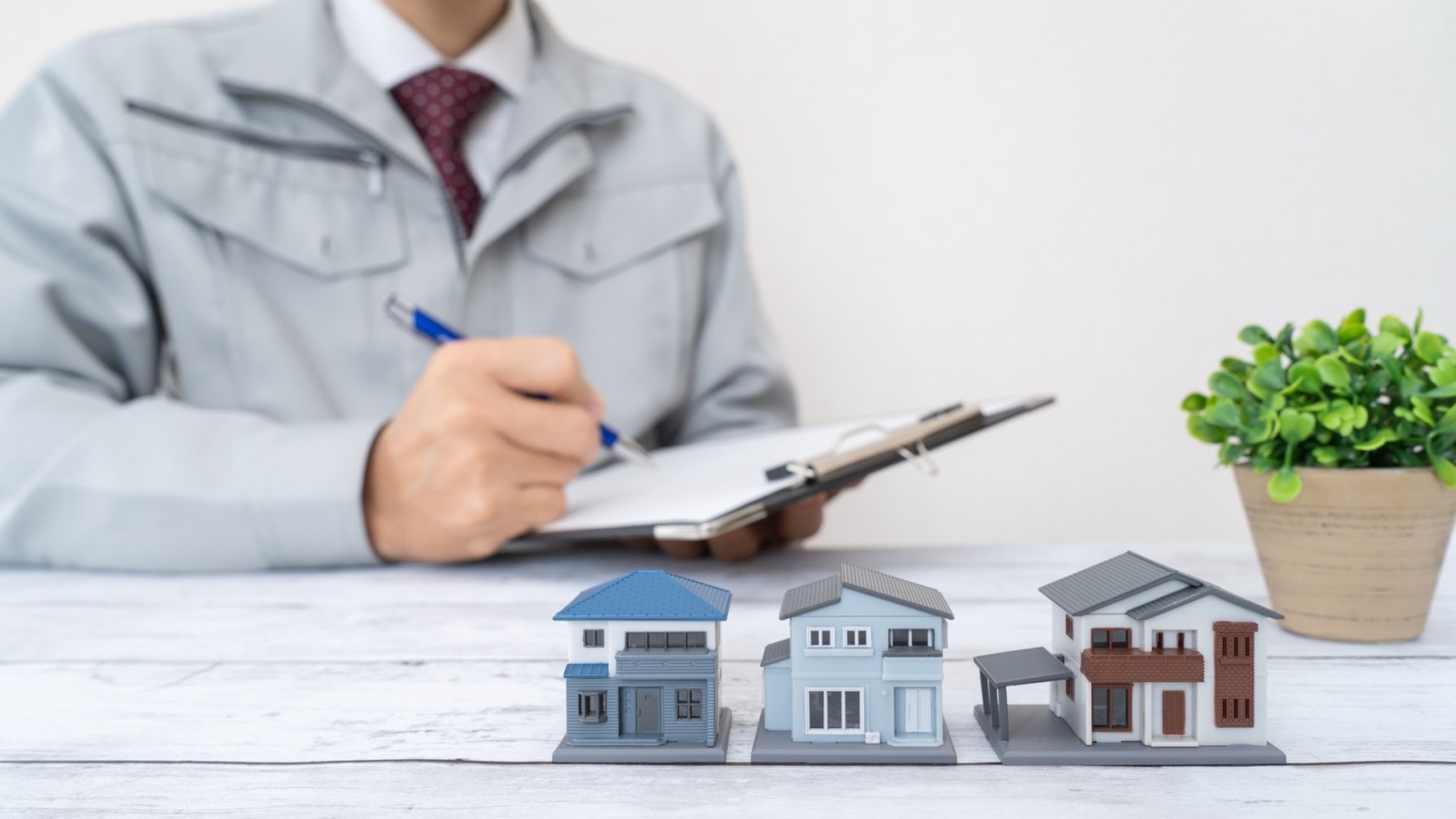 作業着姿の専門家がクリップボードに記入し、手前には3つの異なるデザインの住宅模型が並ぶ。不動産購入の相談、住宅選び、契約手続きのイメージ。