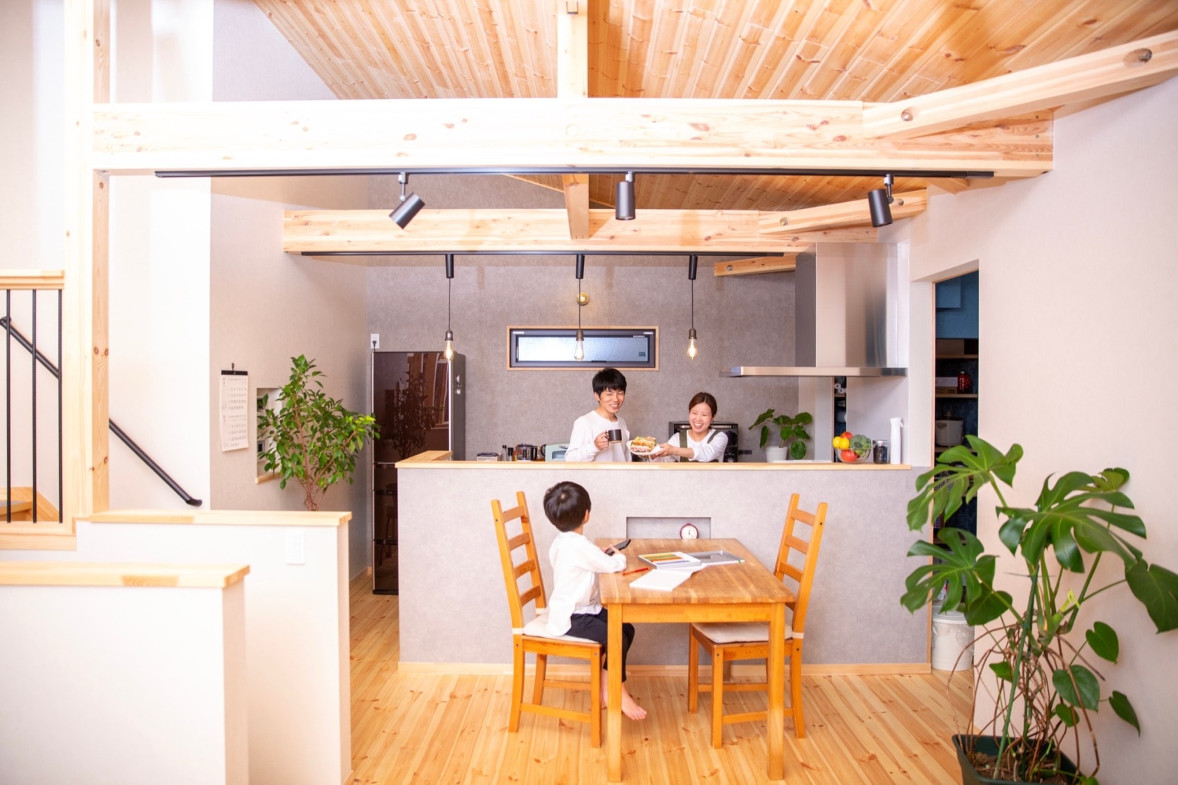 明るい木目の床と天井が開放的なLDKで、キッチンカウンター越しに笑顔の夫婦がダイニングテーブルに座る子供に食事を差し出している。家族の温かい団らんが伝わる、木の温もりを感じるモダンな住宅のイメージ。